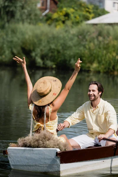 Homem barbudo feliz olhando para a namorada em chapéu de palha com as mãos estendidas durante o passeio de barco — Fotografia de Stock