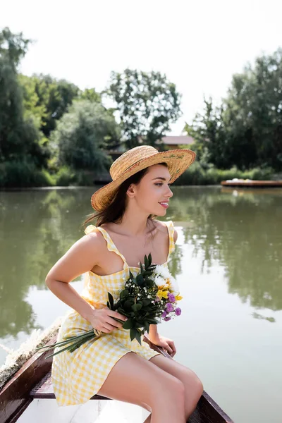 Feliz joven mujer en sombrero de paja y vestido sosteniendo flores mientras que tiene paseo en barco en el lago - foto de stock