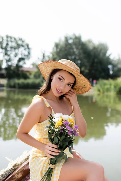 Pretty young woman in straw hat and dress holding flowers while sitting near lake — Stock Photo