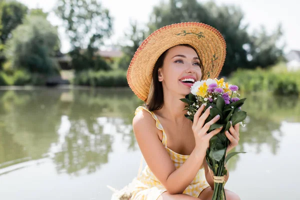 Alegre joven mujer en sombrero de paja y vestido sosteniendo flores cerca del lago - foto de stock