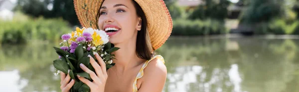 Gioiosa giovane donna in cappello di paglia che tiene fiori vicino al lago, striscione — Foto stock