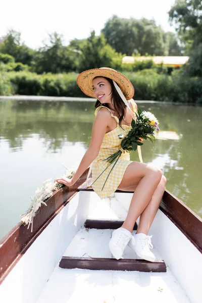Задоволена молода жінка в солом'яному капелюсі і в сукні тримає квіти під час прогулянки на човні на озері — стокове фото