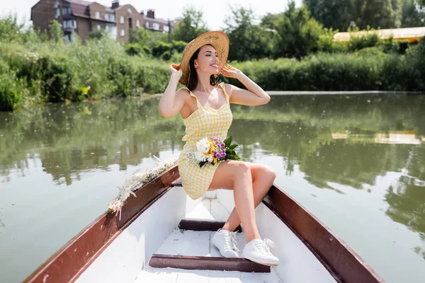 Sorrindo jovem mulher ajustando chapéu de palha e tendo passeio de barco no lago — Fotografia de Stock