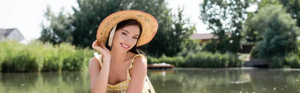 Feliz joven mujer en sombrero de paja mirando a la cámara cerca del río, pancarta - foto de stock