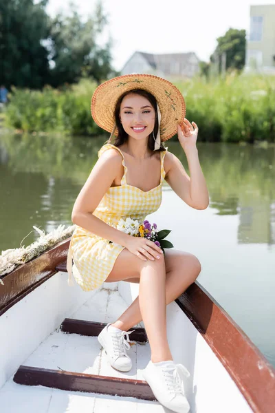 Mujer joven positiva ajustando sombrero de paja y teniendo paseo en barco en el lago - foto de stock