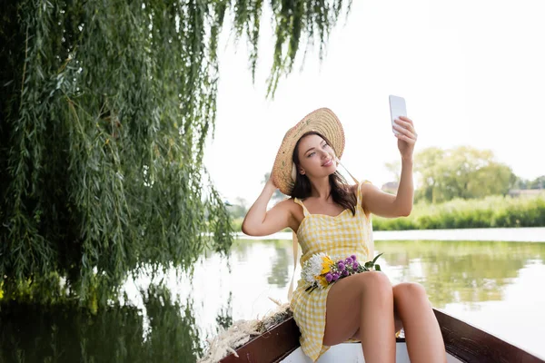 Cheerful young woman in straw hat and dress taking selfie during boat ride on lake — Stock Photo