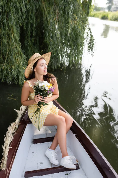 Jeune femme heureuse en chapeau de paille et robe tenant des fleurs pendant la promenade en bateau sur la rivière — Photo de stock