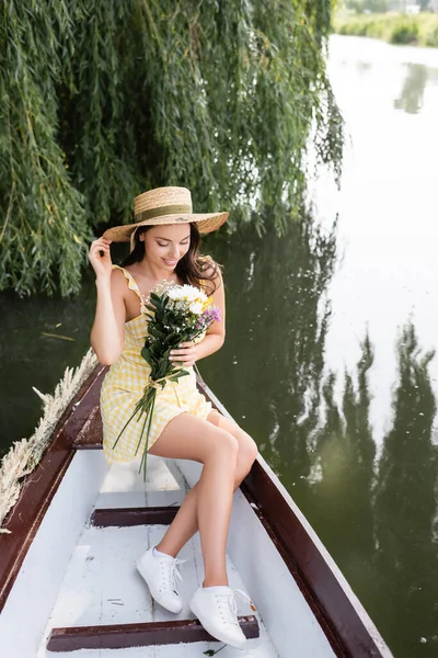 Pleased young woman in straw hat and dress holding flowers during boat ride on river — Stock Photo