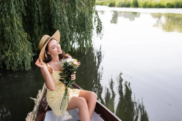 Verträumte junge Frau mit Strohhut und Blumen bei Bootsfahrt auf dem See — Stockfoto