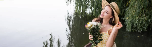 Mujer joven soñadora ajustando sombrero de paja y sosteniendo flores cerca del lago, pancarta - foto de stock