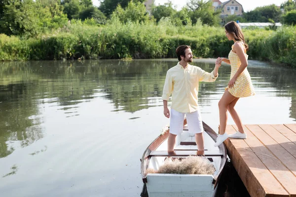 Vista lateral del hombre barbudo sonriente en ropa de verano cogido de la mano con la novia alegre cerca del barco - foto de stock