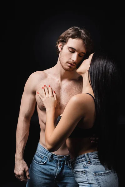 Passionate woman in bra and jeans touching shirtless man isolated on black — Stock Photo