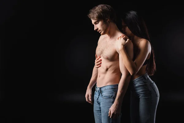 Brunette woman in bra and jeans embracing shirtless man isolated on black — стокове фото