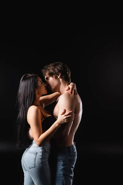 Вид сбоку сексуальной пары в джинсах целующейся и обнимающейся изолированной на черном — стоковое фото