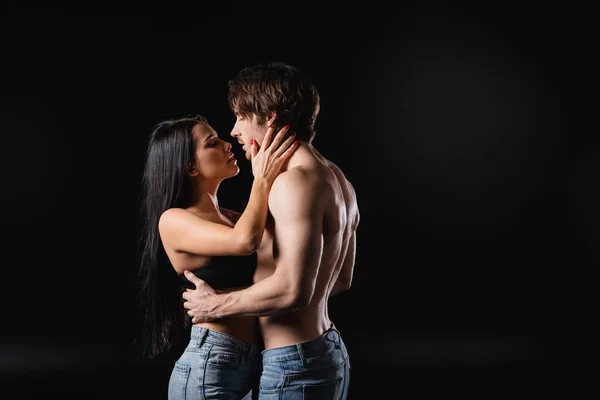 Side view of woman in bra kissing muscular man isolated on black — стоковое фото