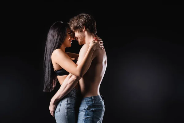 Vue latérale du couple sexy en jeans souriant et câlin isolé sur noir — Photo de stock