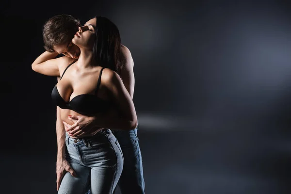 М'язистий чоловік цілує шию спокусливої дівчини в джинсах і бюстгальтер на чорному тлі — стокове фото