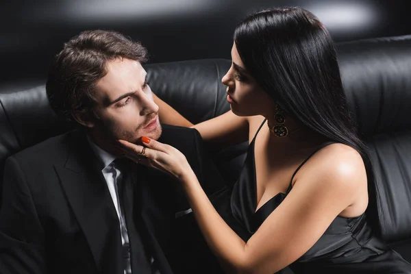 Сексуальна жінка в атласному одязі торкається хлопця в костюмі на дивані на чорному тлі — стокове фото