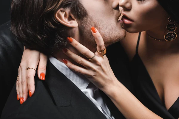 Cropped view of sensual woman kissing boyfriend in suit on couch on black background — стоковое фото