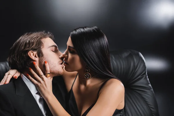 Side view of brunette woman kissing man in formal wear on couch on black background - foto de stock