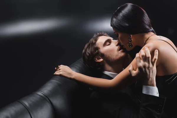 Young man in suit kissing brunette girlfriend on couch on black background — стоковое фото