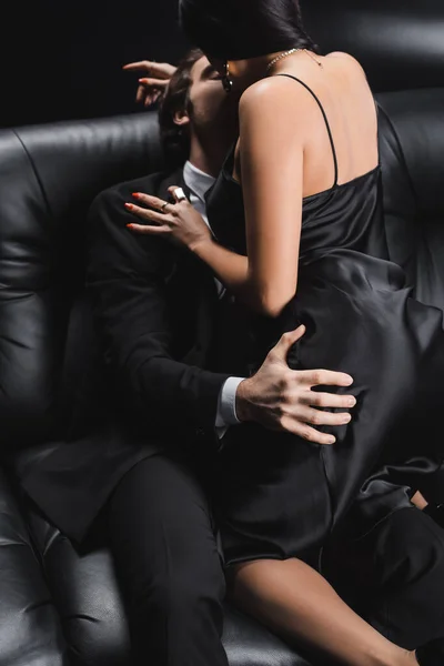 Young man in suit touching buttocks and kissing girlfriend in dress on couch on black background — Fotografia de Stock