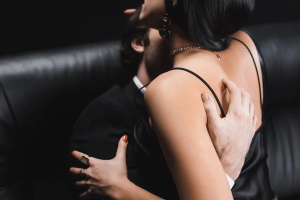 Blurred man touching sexy girlfriend in dress on leather couch isolated on black — Fotografia de Stock