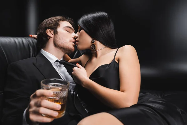 Sensual brunette woman kissing boyfriend in suit holding blurred whiskey on black background — Photo de stock