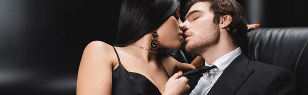 Seductive woman kissing man in suit on leather couch on black background, banner — Stock Photo