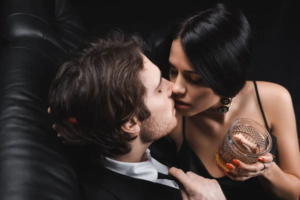 High angle view of brunette woman holding whiskey and kissing boyfriend in suit on couch on black background — Fotografia de Stock