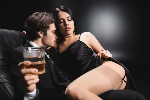 Sensual woman taking off silk dress near boyfriend with blurred glass of whiskey on couch on black background — Foto stock