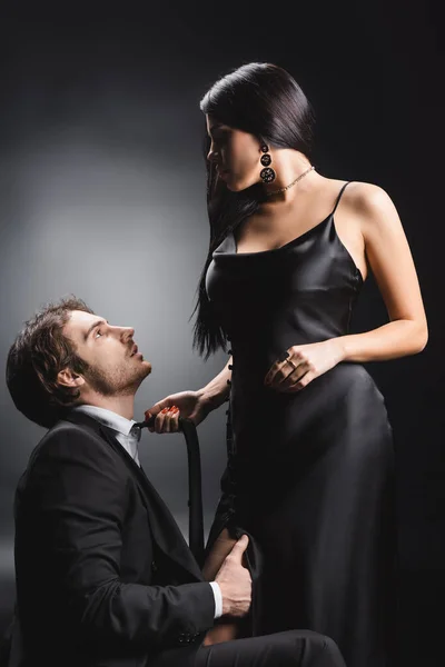 Side view of brunette woman in silk dress holding tie near boyfriend in suit on black background — Fotografia de Stock