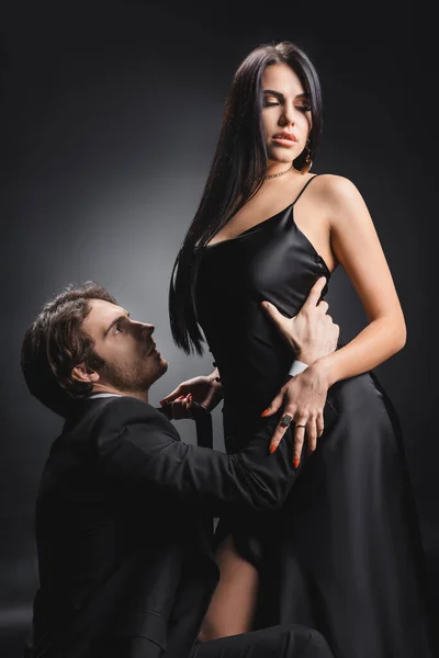 Homme en tenue formelle touchant petite amie sensuelle en robe de soie sur fond noir — Photo de stock