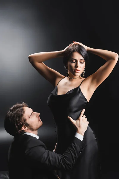 Man in suit touching waist of sexy girlfriend in silk dress on black background — стоковое фото