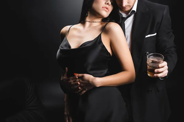 Vista recortada de la sensual mujer en vestido de pie cerca de novio en traje con whisky sobre fondo negro - foto de stock