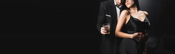 Vista cortada do homem de terno segurando vidro de uísque perto da namorada em vestido de cetim no fundo preto, banner — Fotografia de Stock