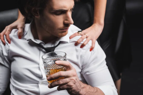 Glass of whiskey in hand of blurred man in shirt sitting near girlfriend on couch isolated on black — Photo de stock