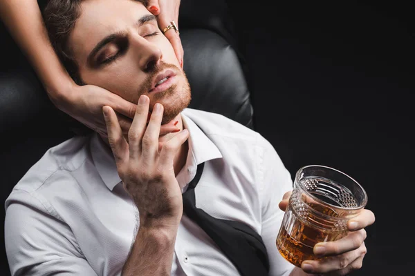 Woman touching face of boyfriend in shirt holding whiskey isolated on black — Stockfoto