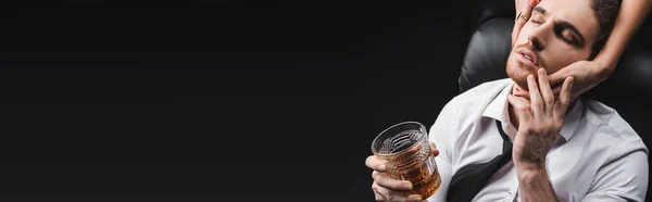 Woman touching face of boyfriend in formal wear holding whiskey isolated on black, banner — Stock Photo