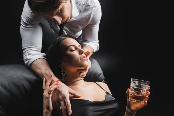 Passionate man touching sexy woman with glass of whiskey on leather couch isolated on black — Fotografia de Stock