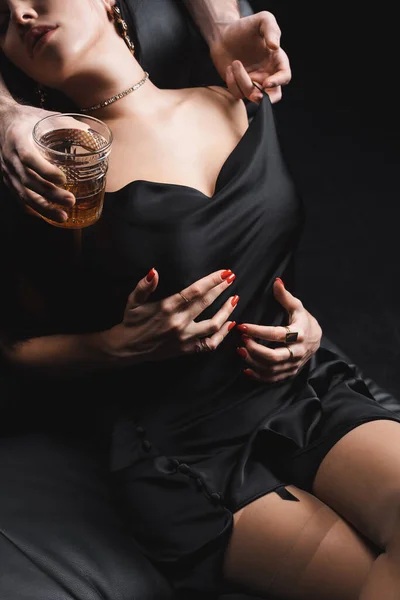 Vista recortada del hombre sosteniendo un vaso de whisky y quitándose el vestido de seda de su novia en el sofá aislado en negro - foto de stock