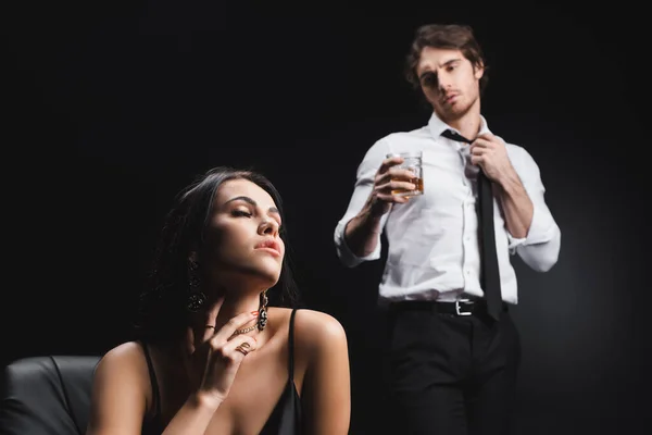 Sensual woman touching neck near blurred boyfriend with glass of whiskey isolated on black — Stockfoto