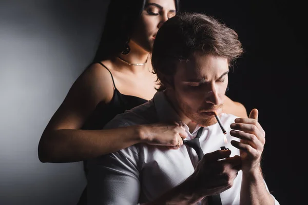 Apasionada mujer desnudando novio en camisa fumar cigarrillo sobre fondo negro - foto de stock