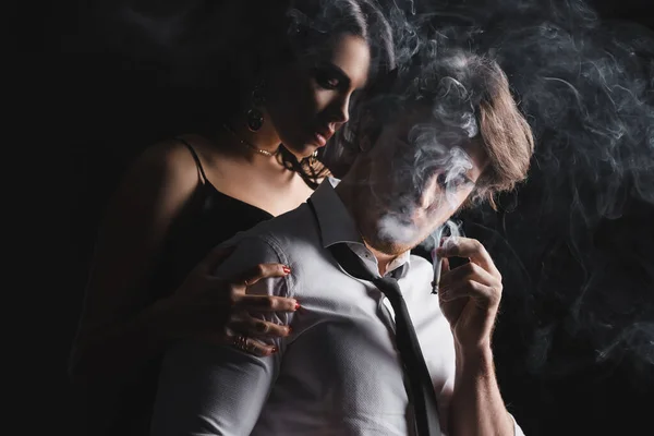 Seductive woman hugging man in formal wear with cigarette on black background — Stockfoto