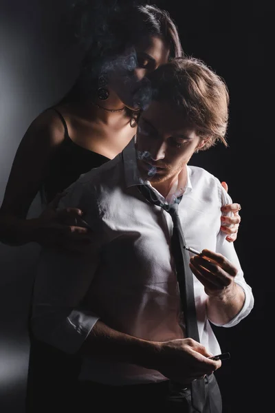 Woman in dress kissing boyfriend in formal wear holding cigarette on black background — Stock Photo