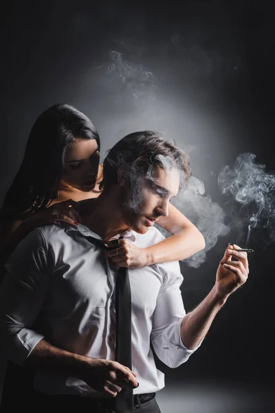 Young man in formal wear holding cigarette near sexual girlfriend on black background — Stockfoto