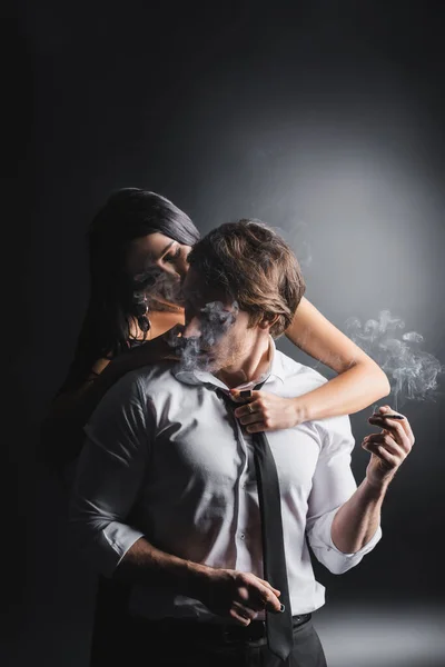 Sexy woman touching tie on boyfriend holding cigarette on black background — Foto stock