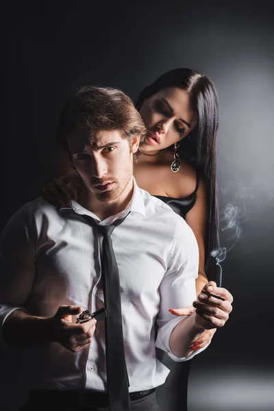 Uomo in abbigliamento formale in possesso di sigaretta e guardando la fotocamera vicino donna sexy su sfondo nero — Foto stock