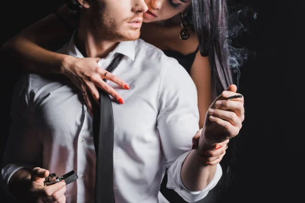 Cropped view of man holding cigarette and lighter near blurred sexy girlfriend on black background — Stock Photo