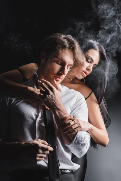 Sexy brunette woman touching boyfriend with cigarette near smoke on black background — Stock Photo